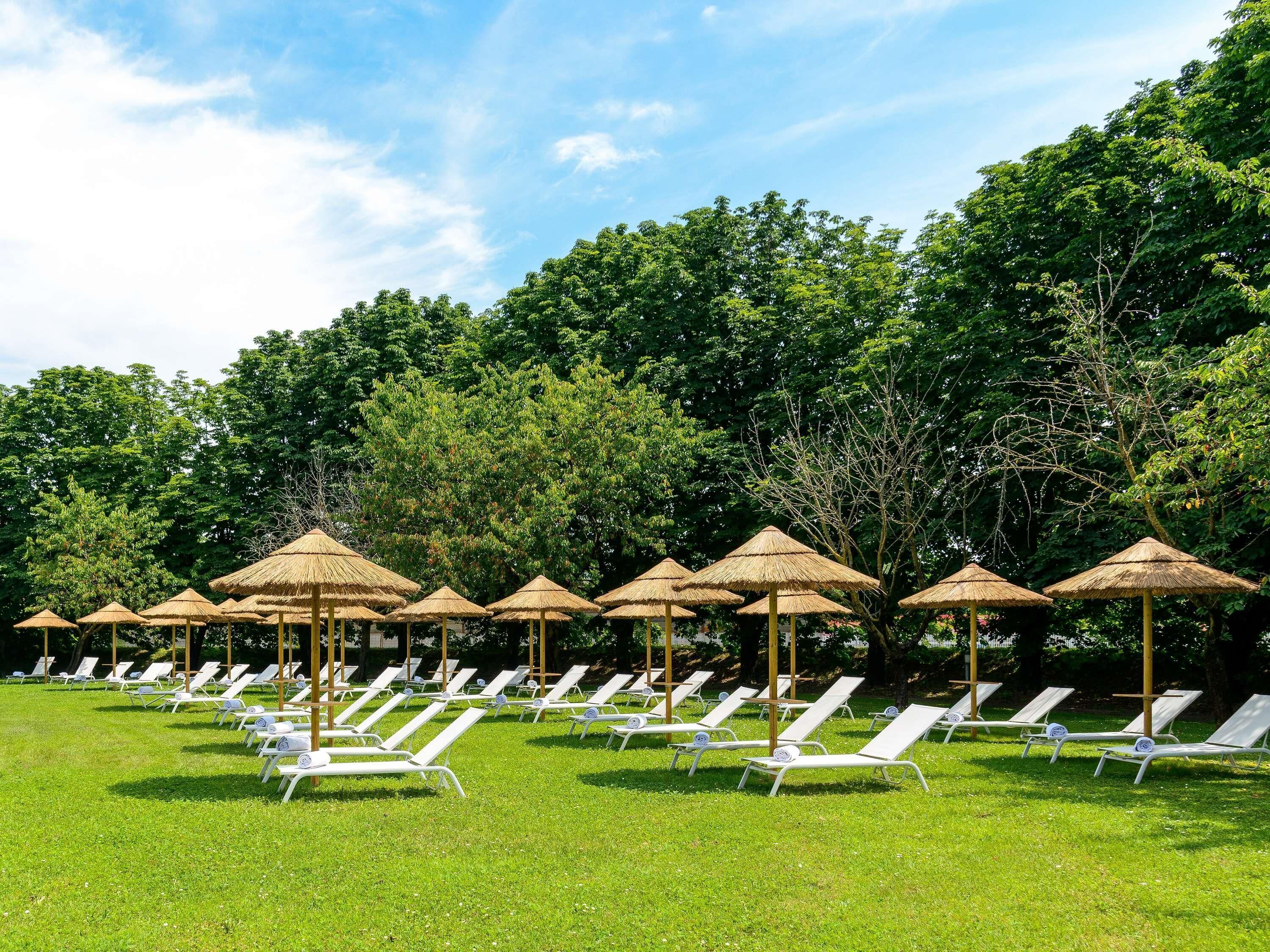 Een rij witte loungestoelen met rieten parasols, op een gazon met bomen op de achtergrond.