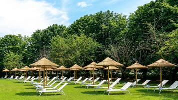 Een rij witte loungestoelen met rieten parasols, op een gazon met bomen op de achtergrond.