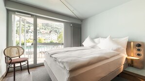 Ein Hotelzimmer mit einem großen Bett, einem Sessel, einem Nachttisch mit Lampe und Blick auf Gebäude durch ein großes Fenster.