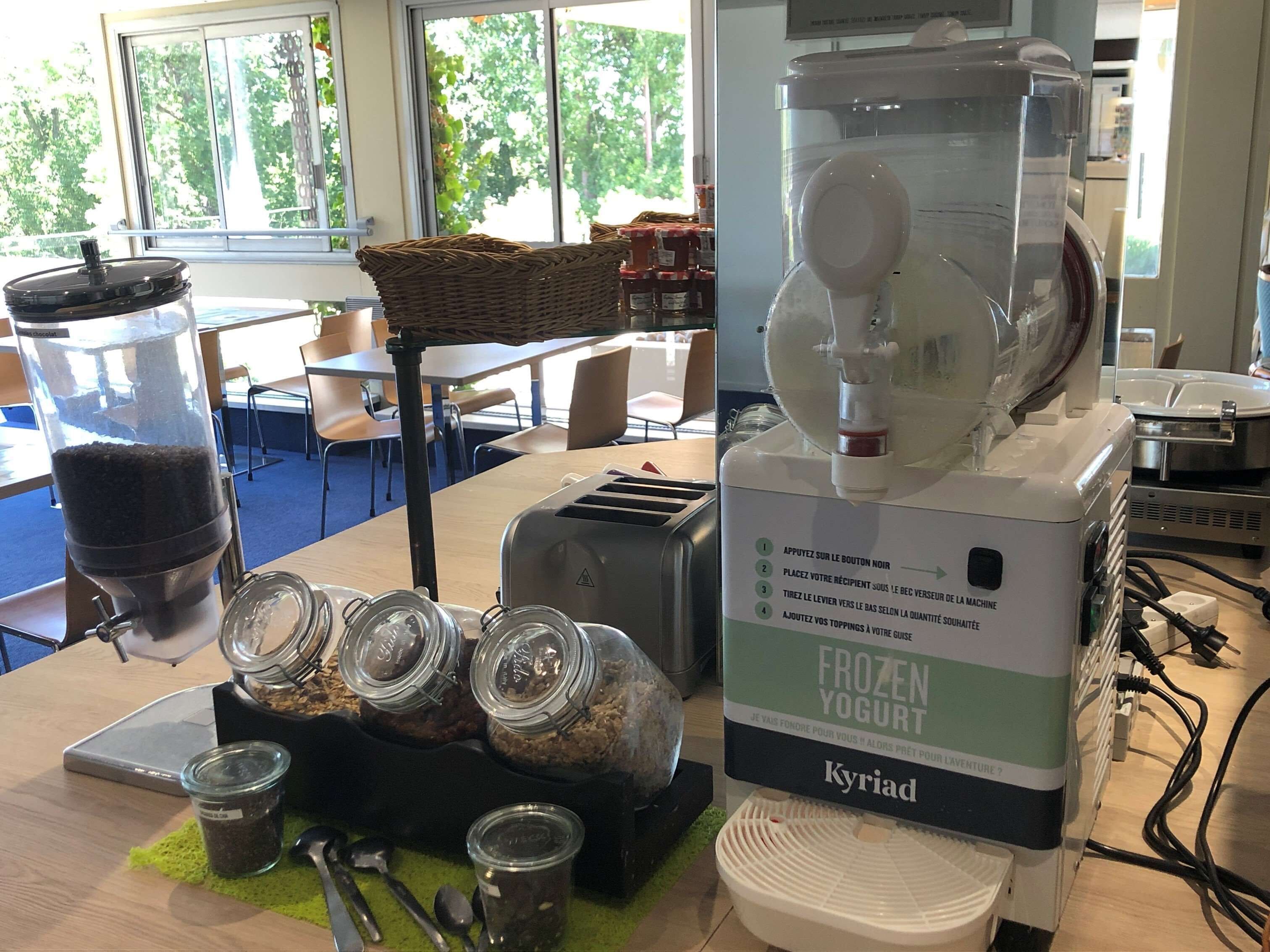 Une machine à café et un distributeur de yaourt glacé sont posés sur une table, à côté de pots de café et d’ustensiles.