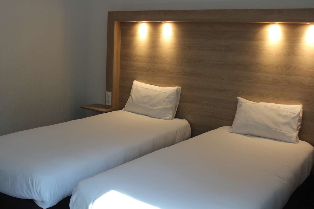 A hotel room with two single beds, wooden headboard, bedside tables, and wall-mounted lights.