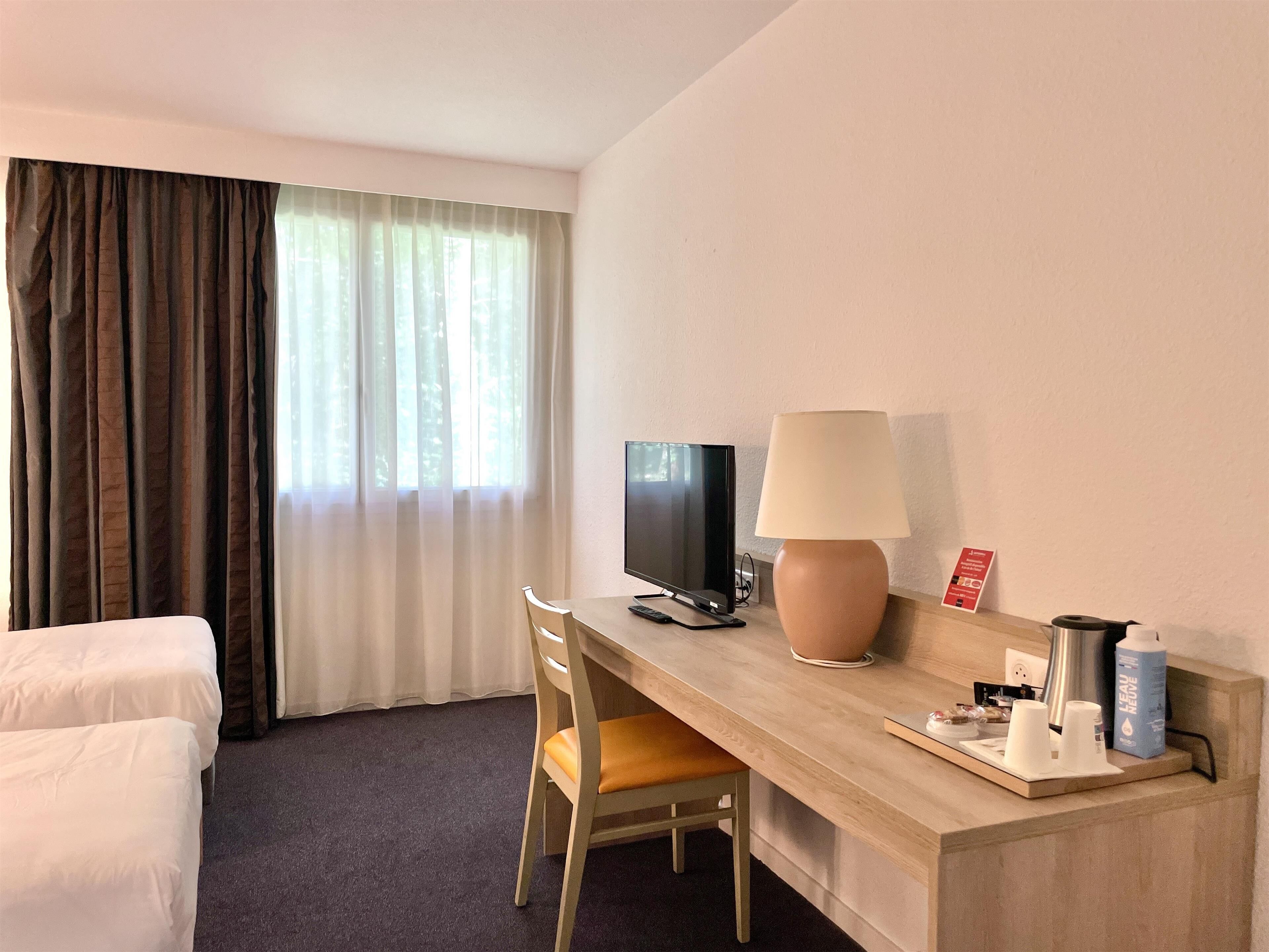 A hotel room with a bed, a desk with a lamp, a chair, a television, and a window with curtains.