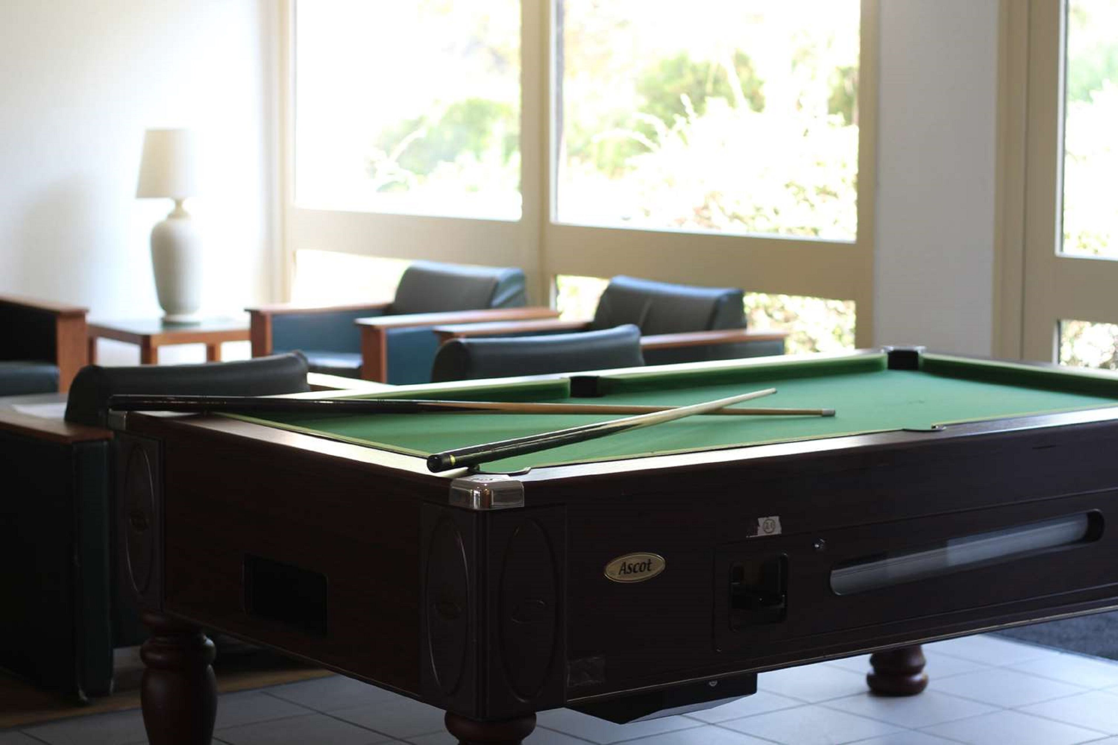 A pool table with a green felt surface, a cue stick, and a pool table branded "Ascot."