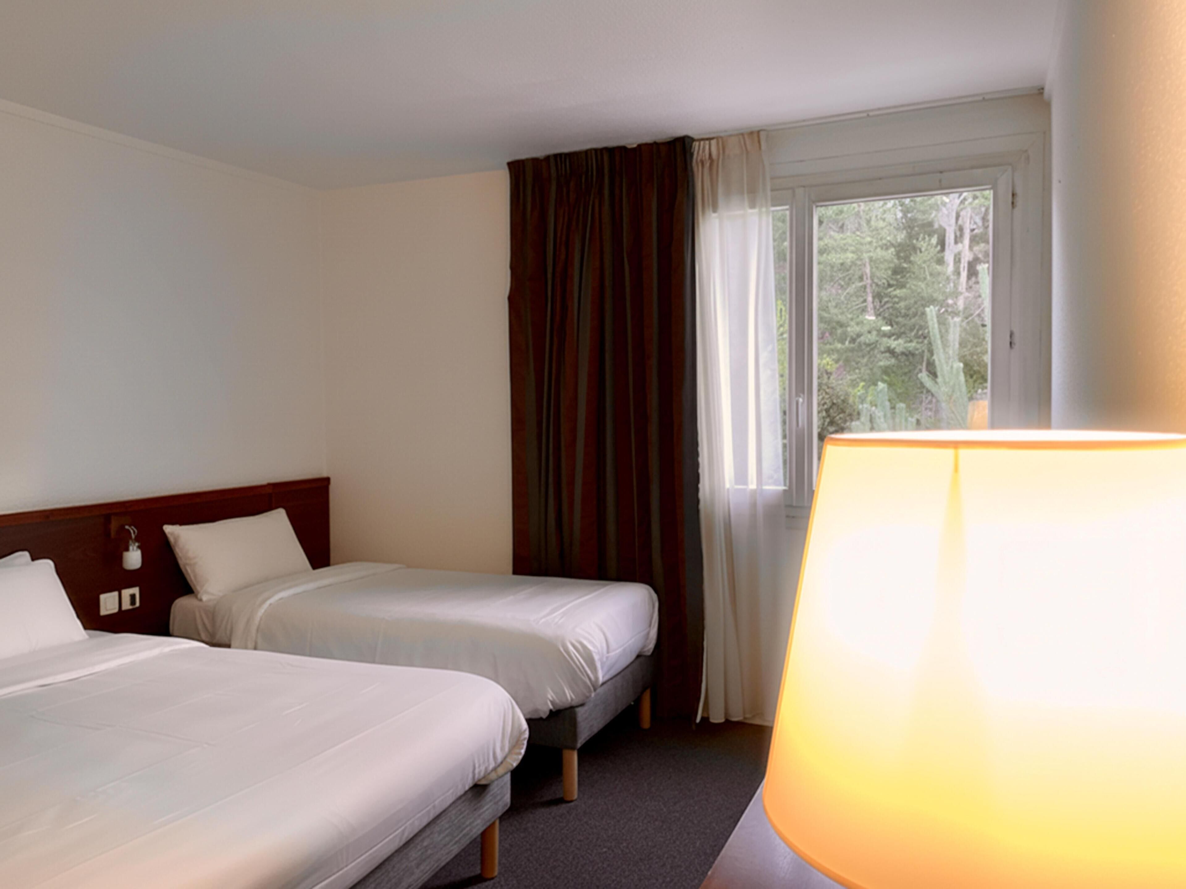 Une chambre d’hôtel avec deux lits simples, une fenêtre avec des rideaux et une lampe sur la table de chevet.