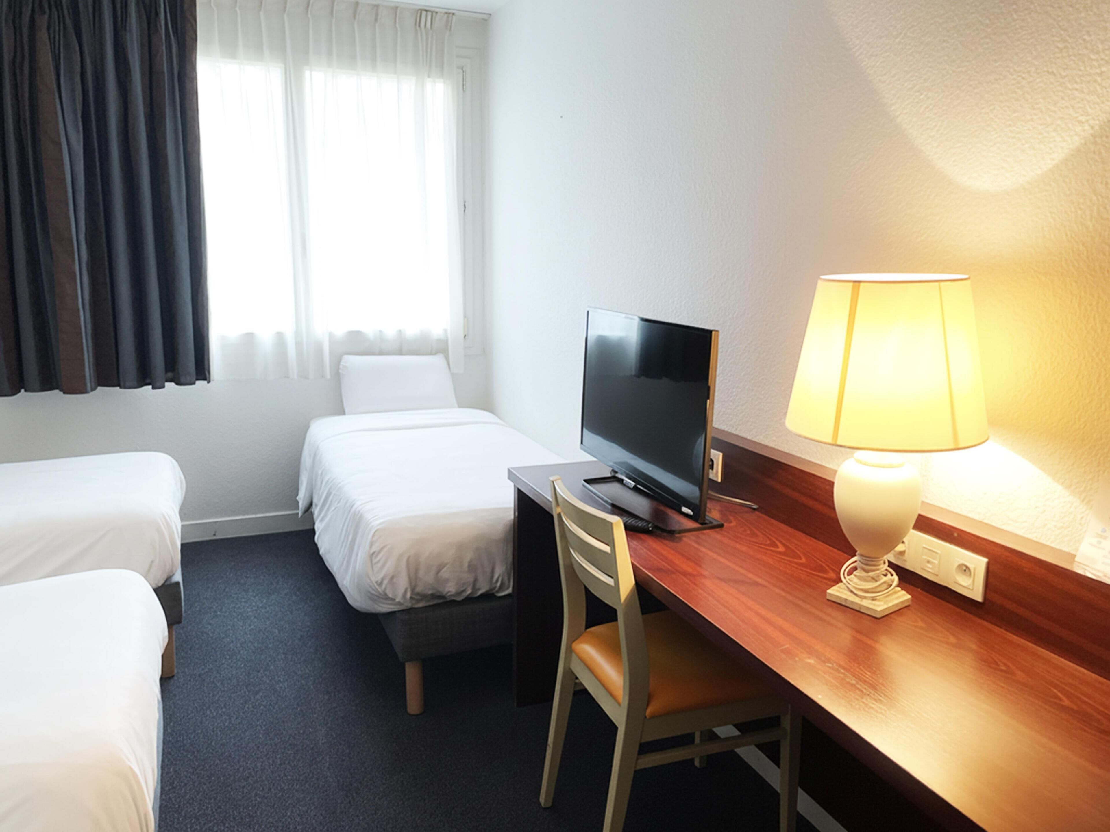 A hotel room with two single beds, a desk with a television, a chair, and a bedside lamp.