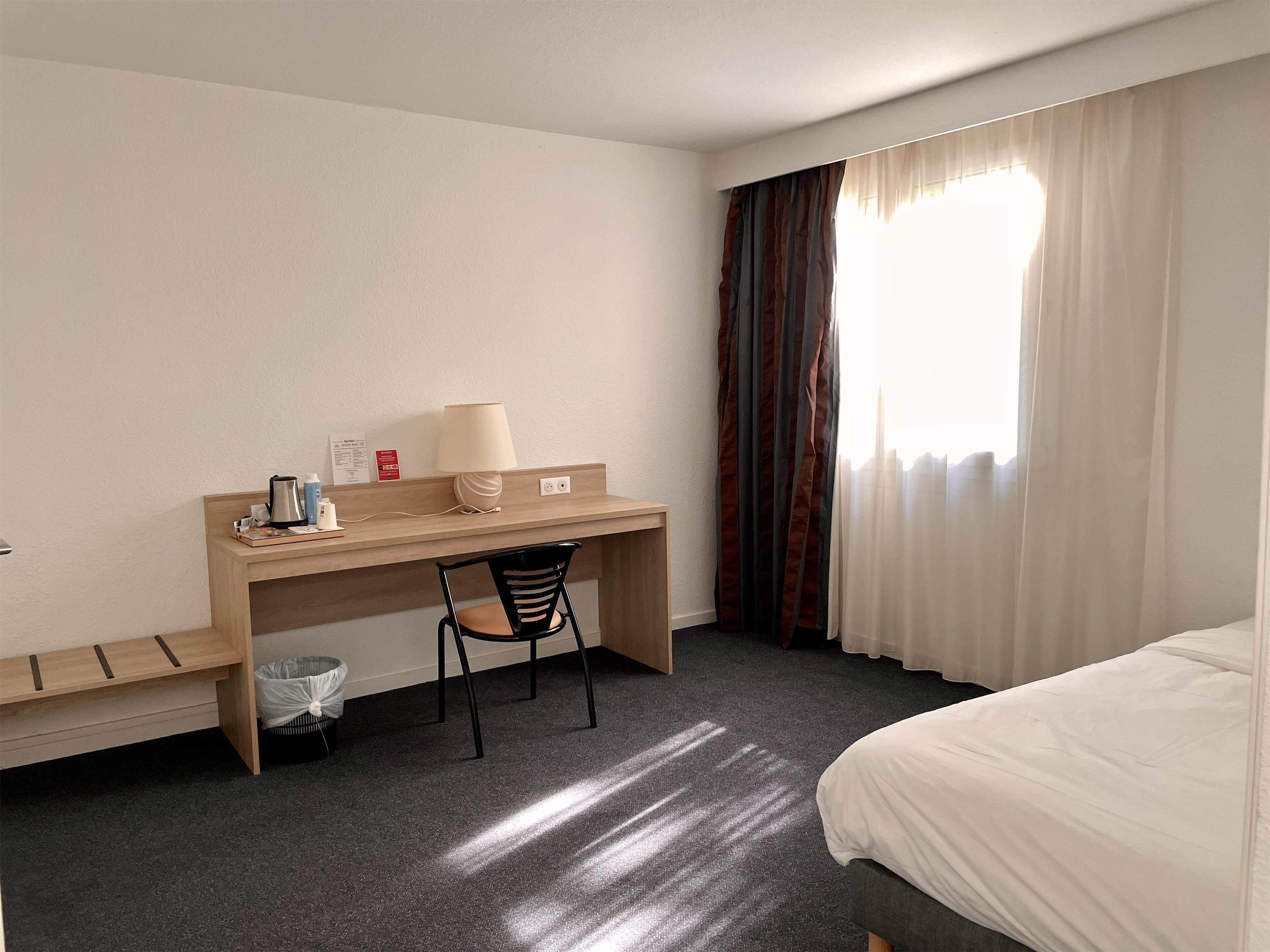 A hotel room with a bed, a desk with a chair, a lamp, and a window with curtains.