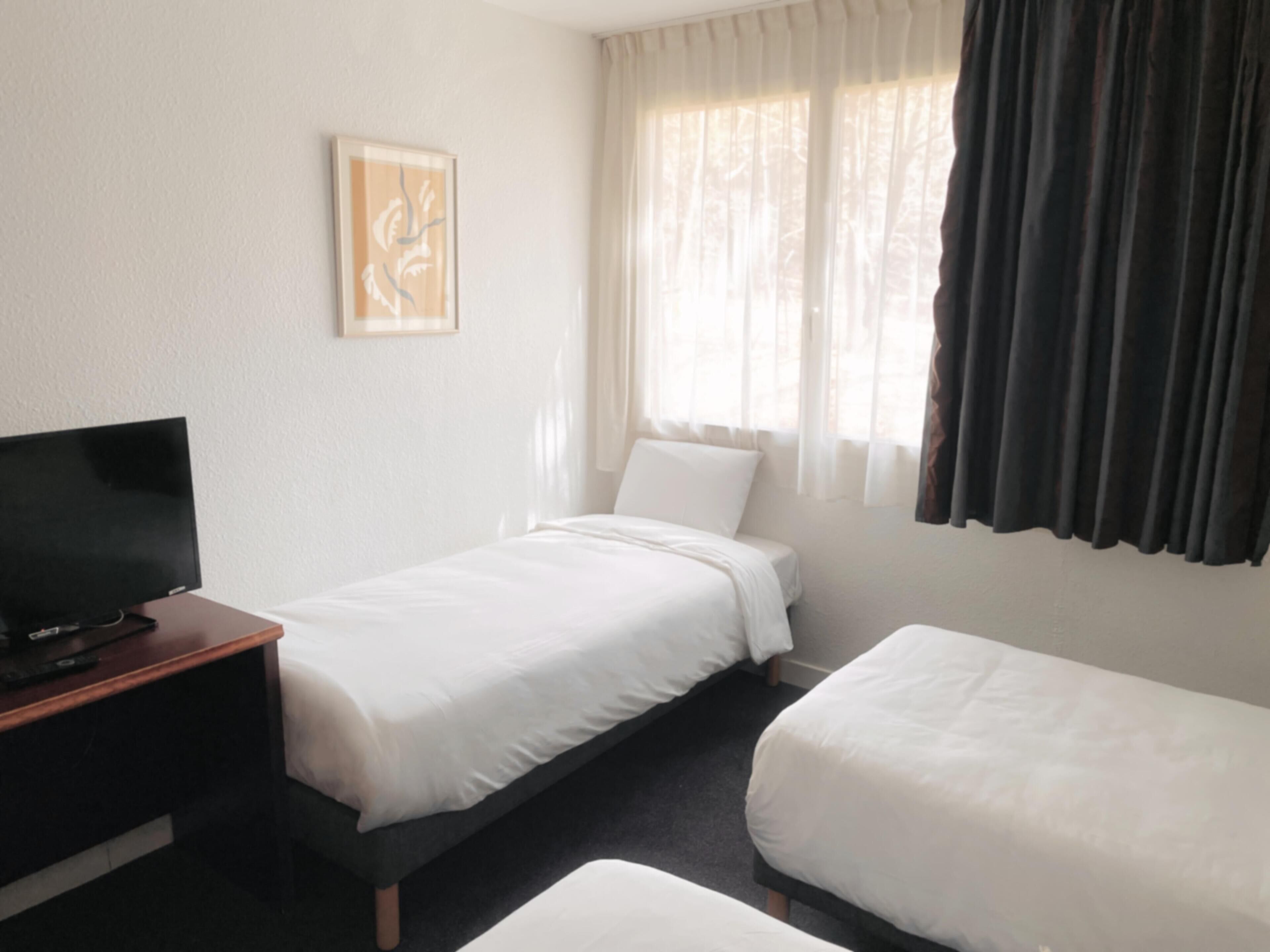 Een hotelkamer met twee bedden, een televisie op een houten standaard en een raam met gordijnen.