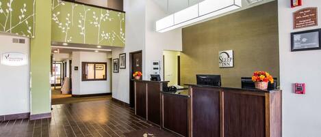A hotel reception area with a wooden reception desk, a 'Sleep Inn' sign, a fire alarm, and a 'Welcome' mat.