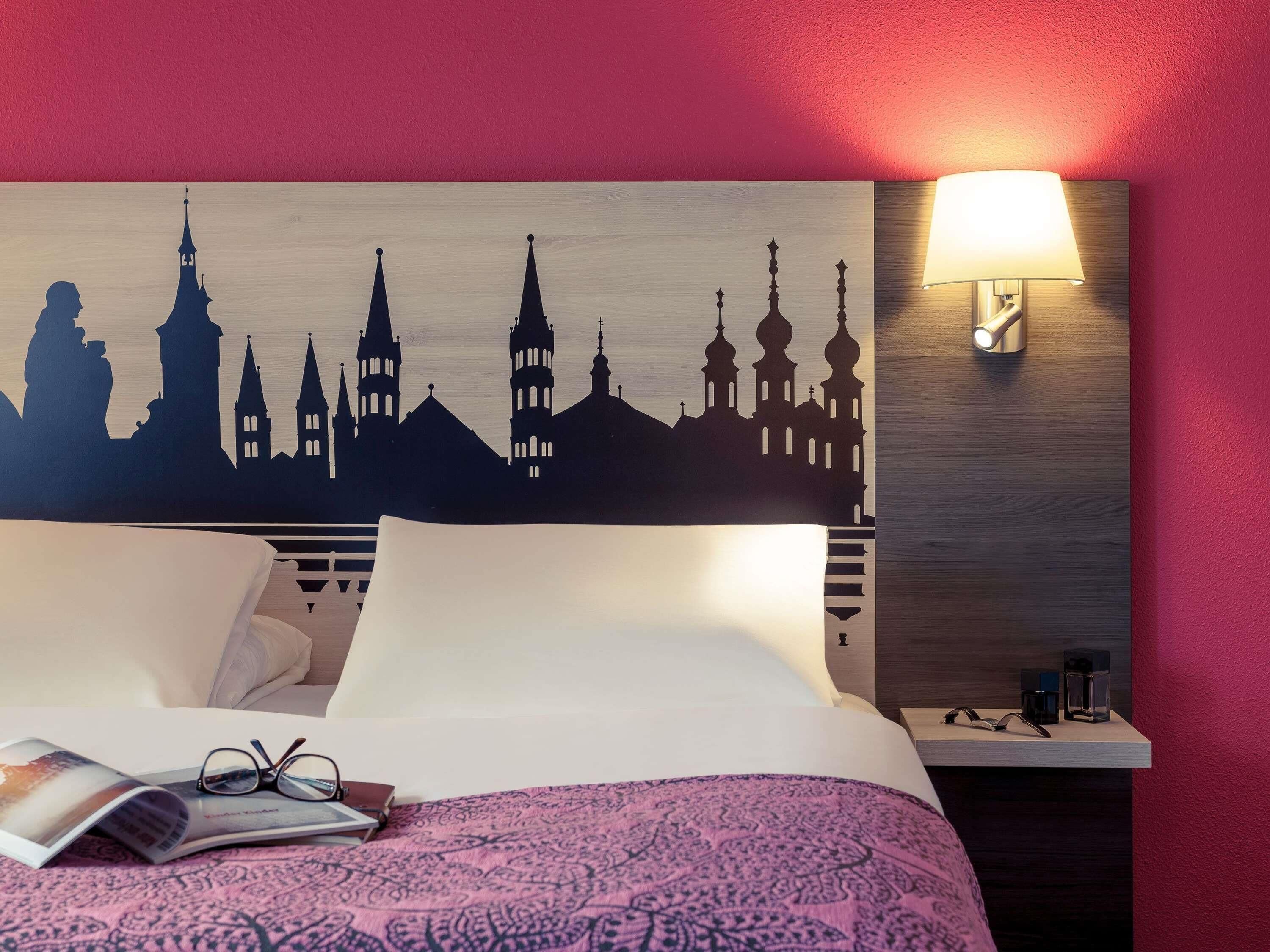 A hotel room with a bed, a nightstand, a lamp, and a wall decoration depicting European architecture.