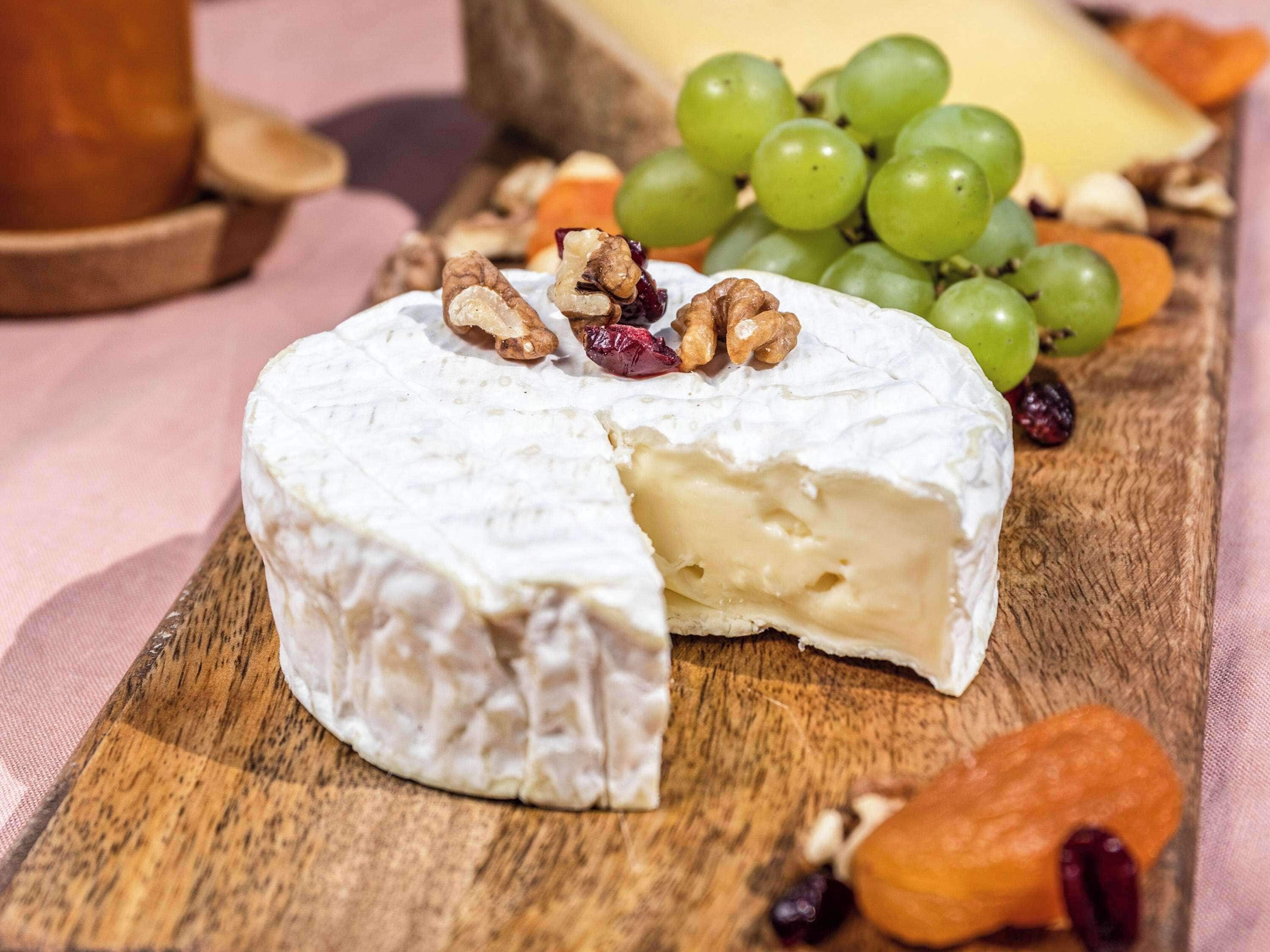 Une portion de fromage, dont on a déjà coupé une tranche, est présentée sur une planche en bois, entourée de raisins, de noix et de fruits secs.