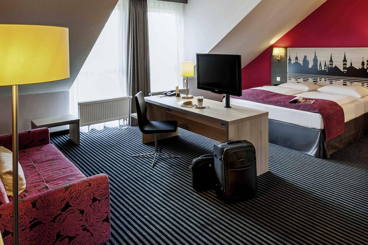 A hotel room with a desk, two beds, a TV, a lamp, a couch, and a suitcase.