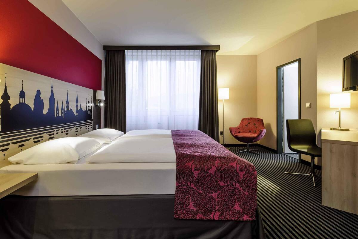 A hotel room with a bed, a chair, a desk, and a lamp.
