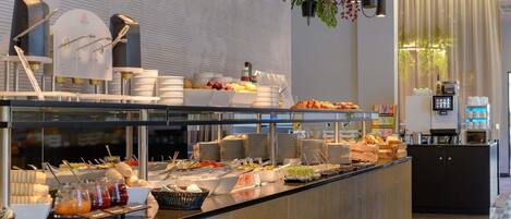 Zona de bufé con diversos alimentos, una máquina de café y plantas colgantes.