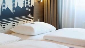 A hotel room with a bed, white pillows, a wall-mounted lamp, and a window with curtains.