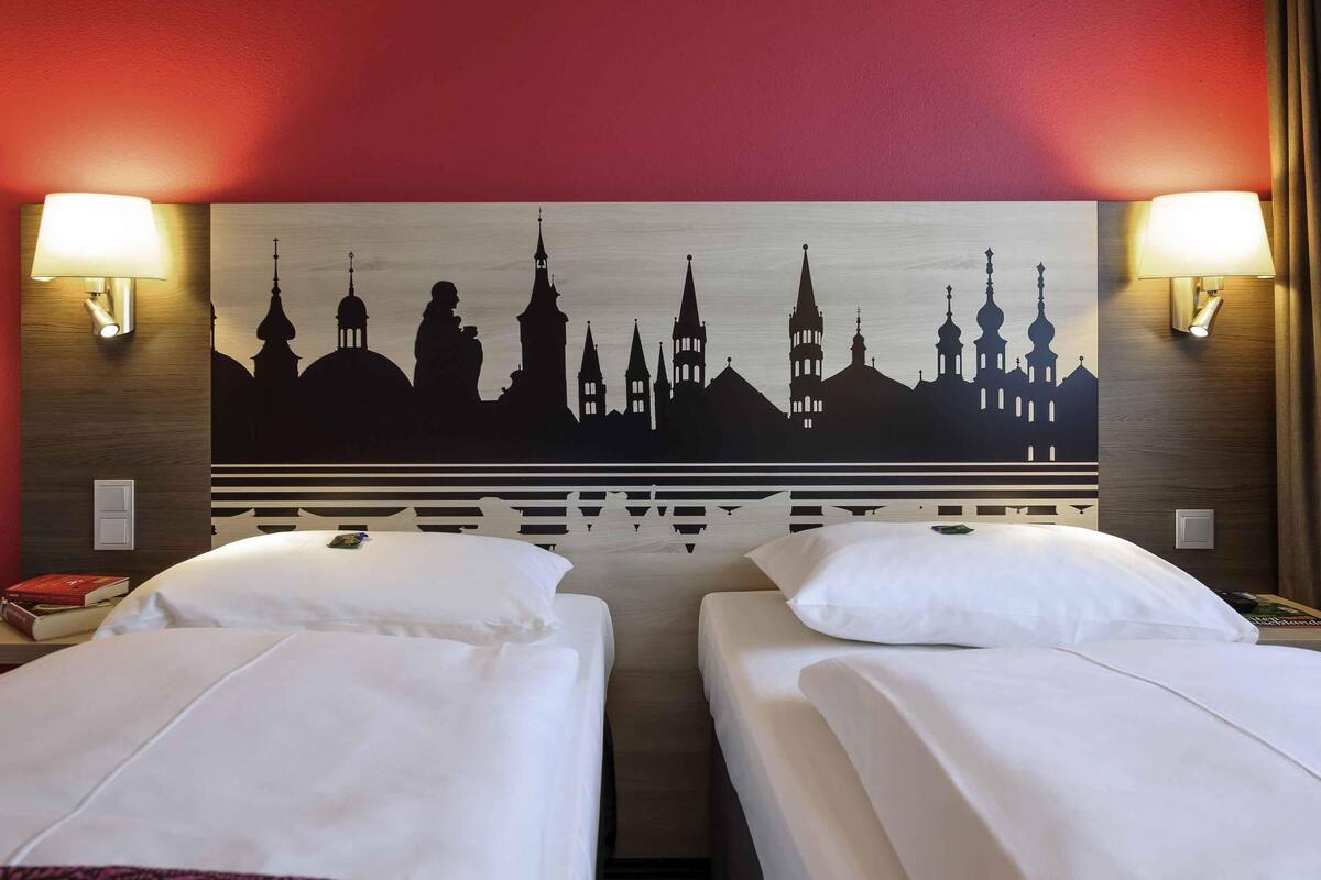 A hotel room with two beds, a city skyline mural on the headboard, and wall-mounted lamps.