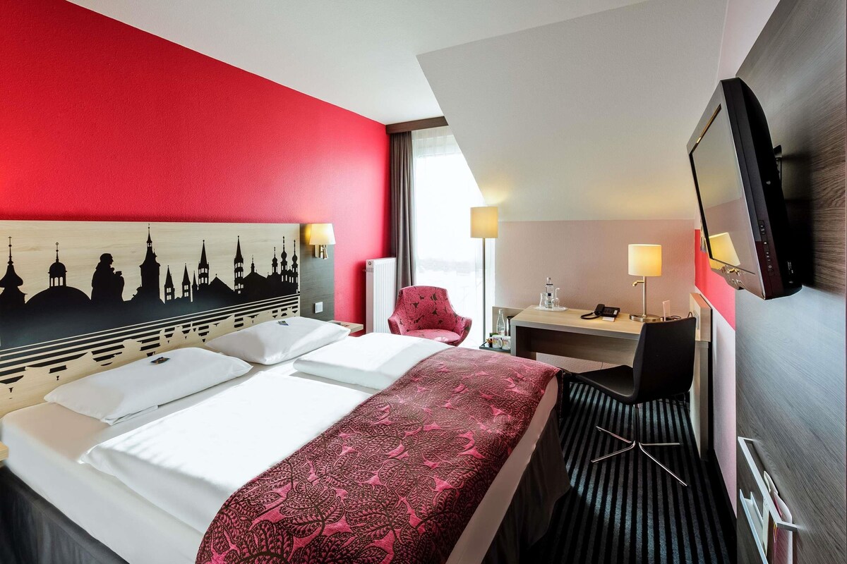 A hotel room with a bed, desk, chair, and a TV.