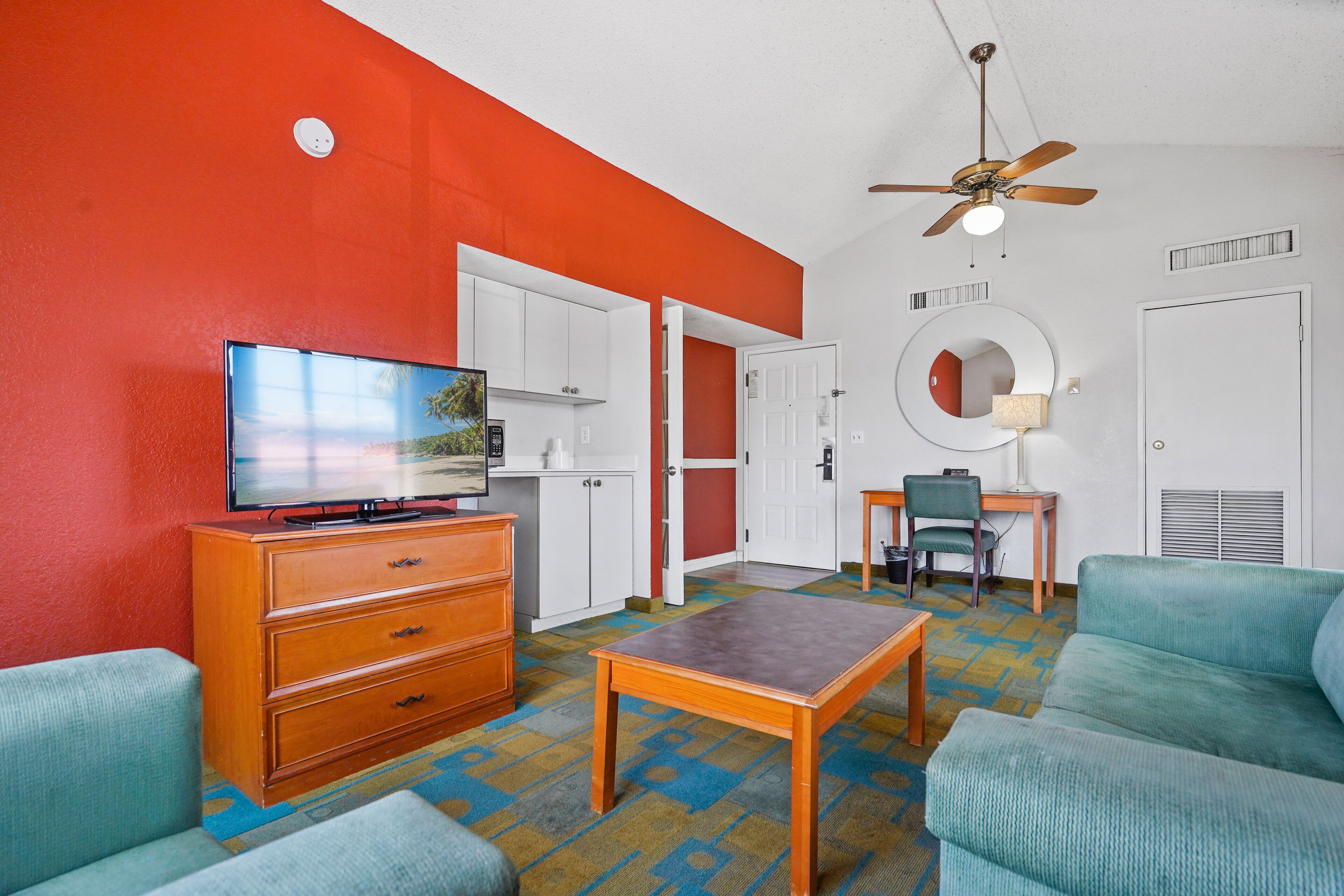 A room with a television, wooden dresser, teal sofa, and a ceiling fan.