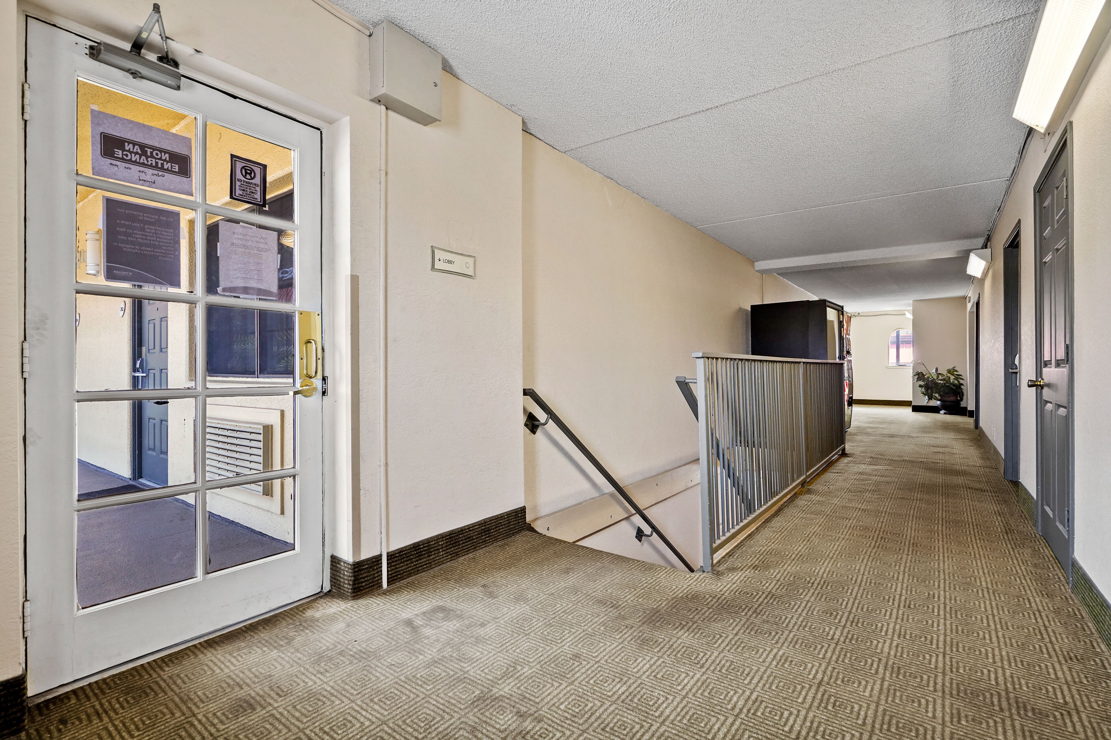 A hotel corridor with a patterned carpet, a glass door with multiple panes, a sign indicating a lobby, and a metal railing along the stairs.