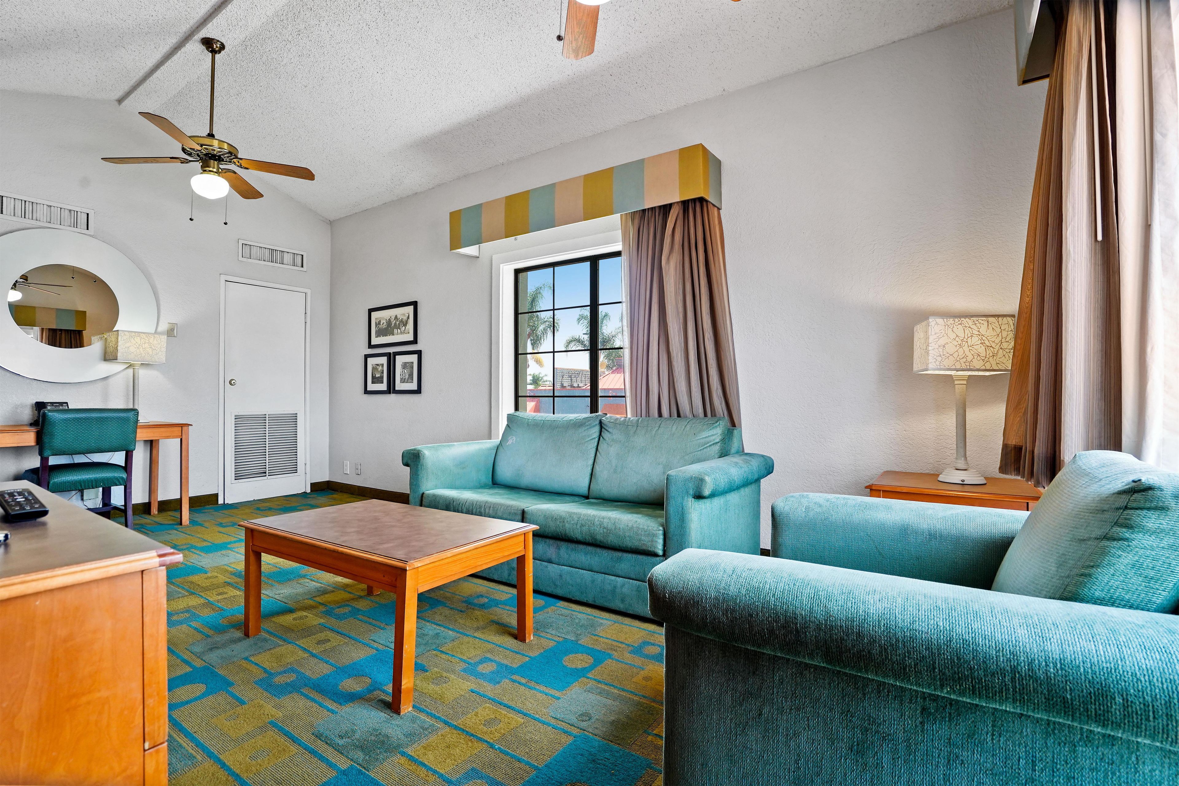 A hotel room with a sofa, armchairs, a coffee table, a desk, a chair, a lamp, a ceiling fan, a window with curtains, and a wall-mounted mirror.