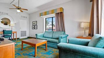 A hotel room with a sofa, armchairs, a coffee table, a desk, a chair, a lamp, a ceiling fan, a window with curtains, and a wall-mounted mirror.