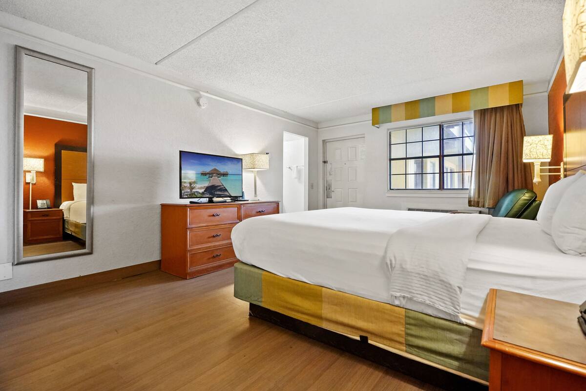 A hotel room with a large bed, wooden floor, a dresser, a painting, a window with curtains, and a door.