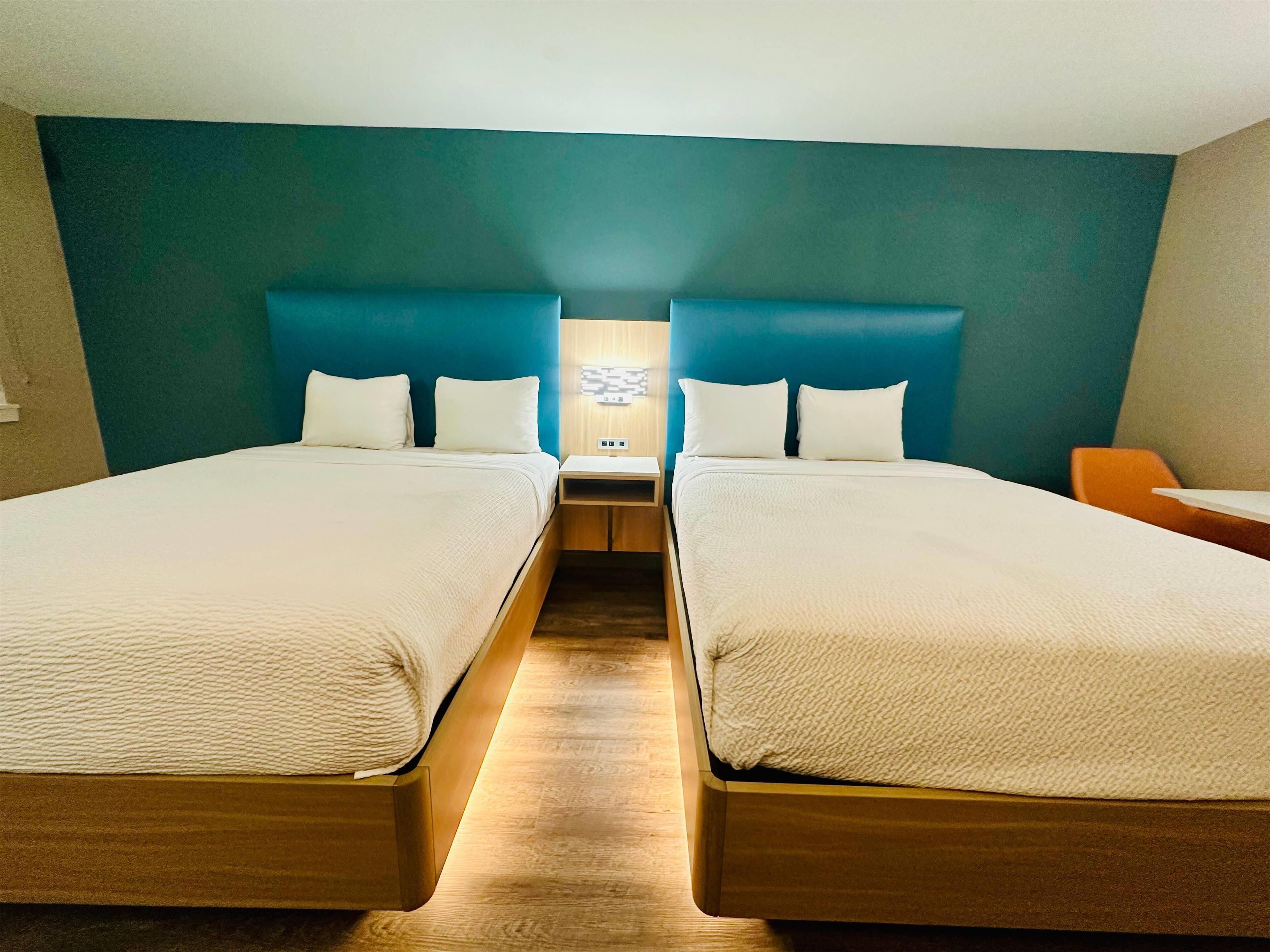 A hotel room with two single beds, a wooden floor, teal walls, and a nightstand with a lamp.