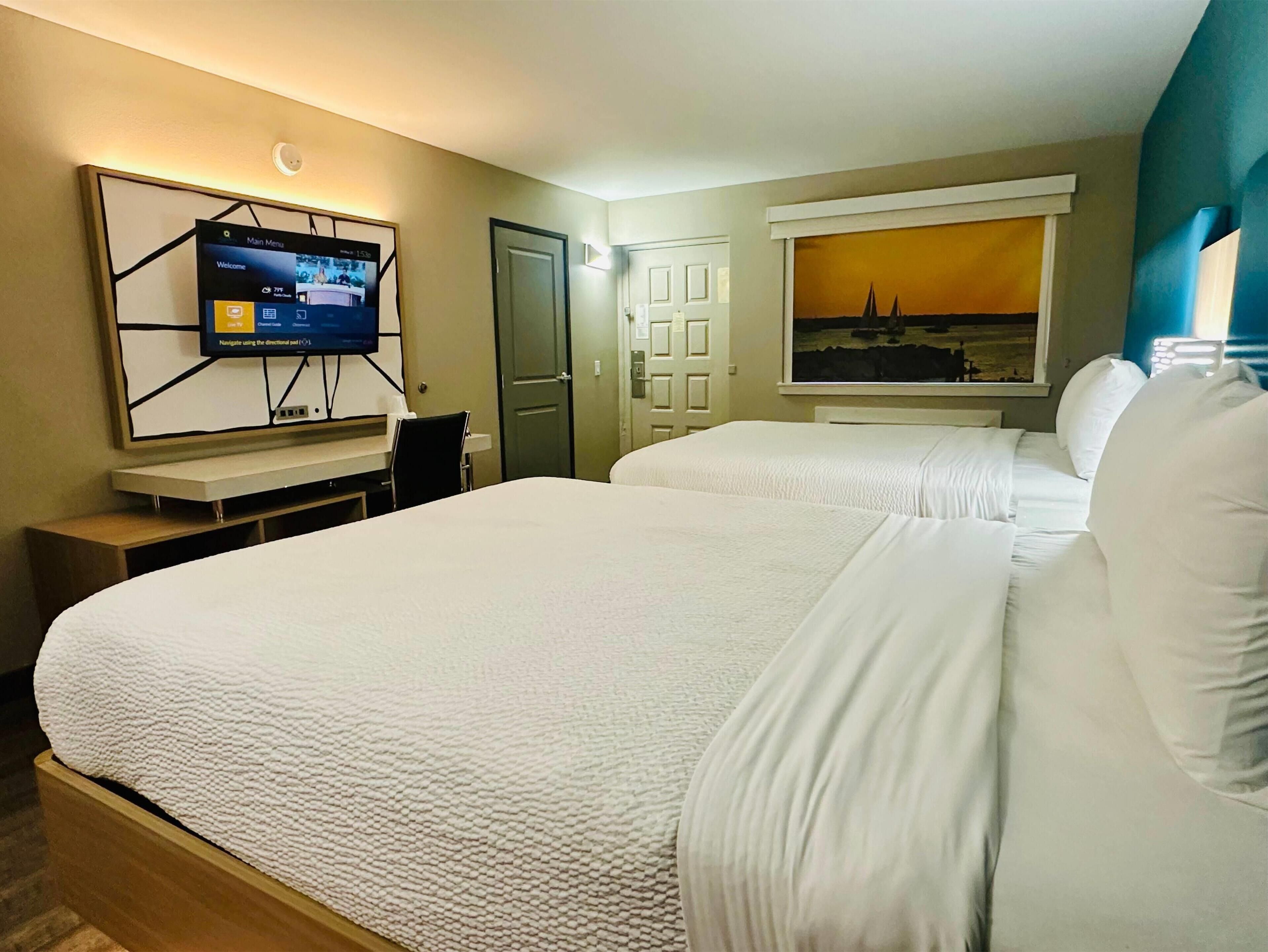 Habitación de hotel con dos camas, un televisor montado en la pared, una puerta y una ventana con un cuadro.