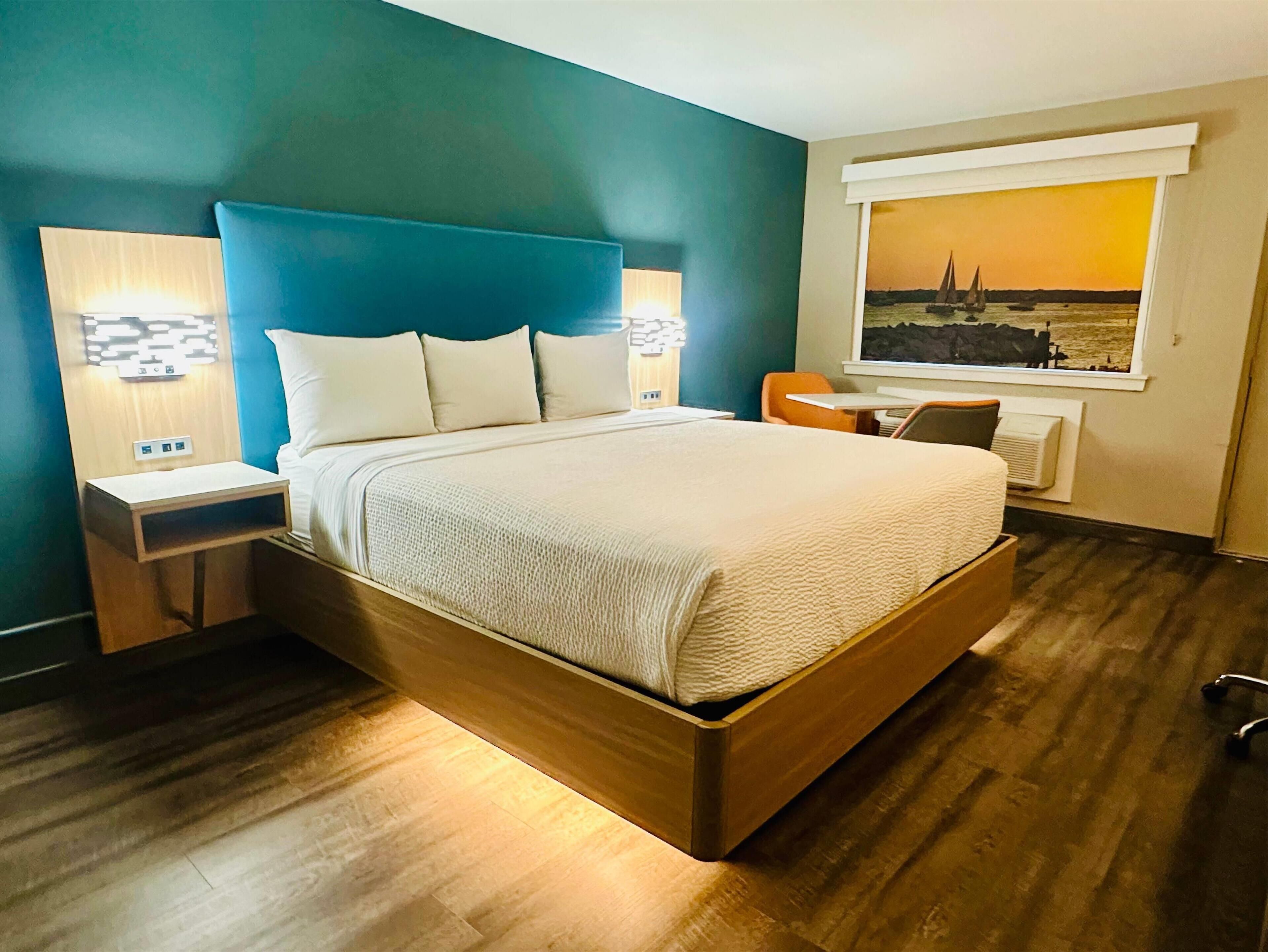 Una habitación de hotel moderna con una cama grande, dos mesitas de noche con lámparas, una pequeña mesa, una silla y una ventana con vistas panorámicas.