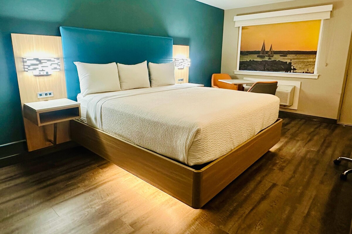 A modern hotel room with a large bed, two bedside tables with lamps, a small table, a chair, and a window with a scenic view.