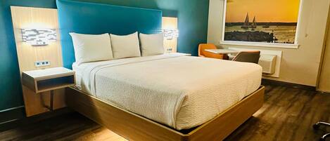 Una habitación de hotel moderna con una cama grande, dos mesitas de noche con lámparas, una pequeña mesa, una silla y una ventana con vistas panorámicas.