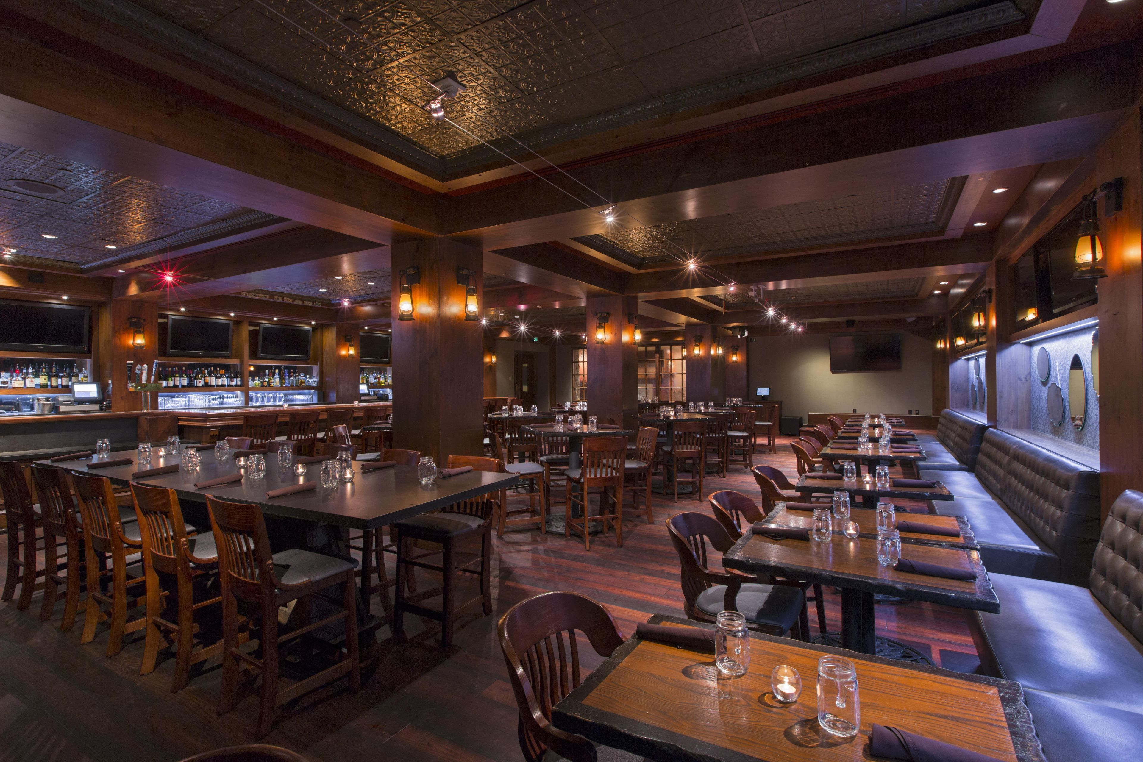 A restaurant with multiple dining tables and chairs, a bar area with bottles, and a high ceiling with decorative lighting.