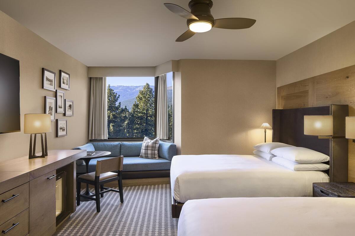 A hotel room with a desk, two beds, a sofa, and a large window with a mountain view.