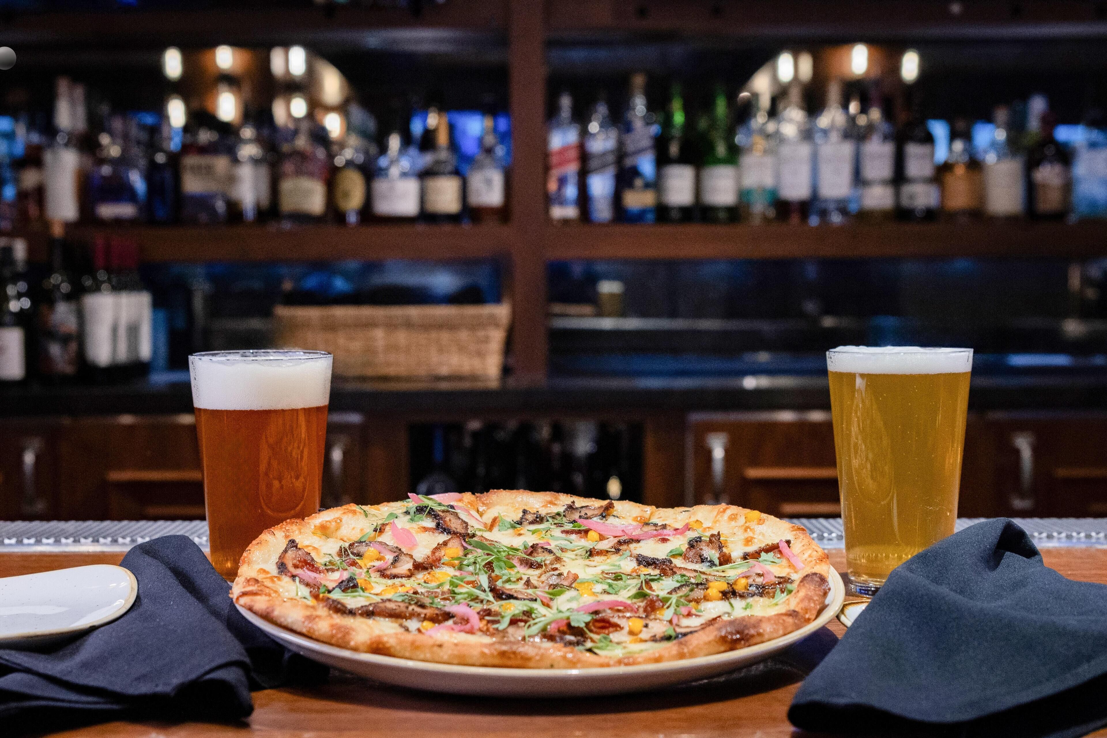 Une pizza garnie de divers ingrédients et deux verres de bière, le tout posé sur une table dans un bar.