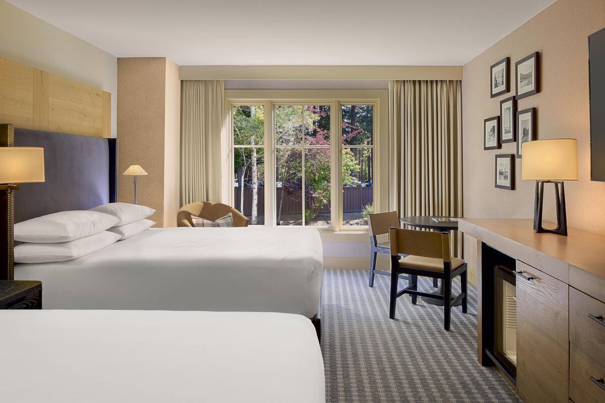 A hotel room with two beds, a desk, a chair, and a large window with curtains.