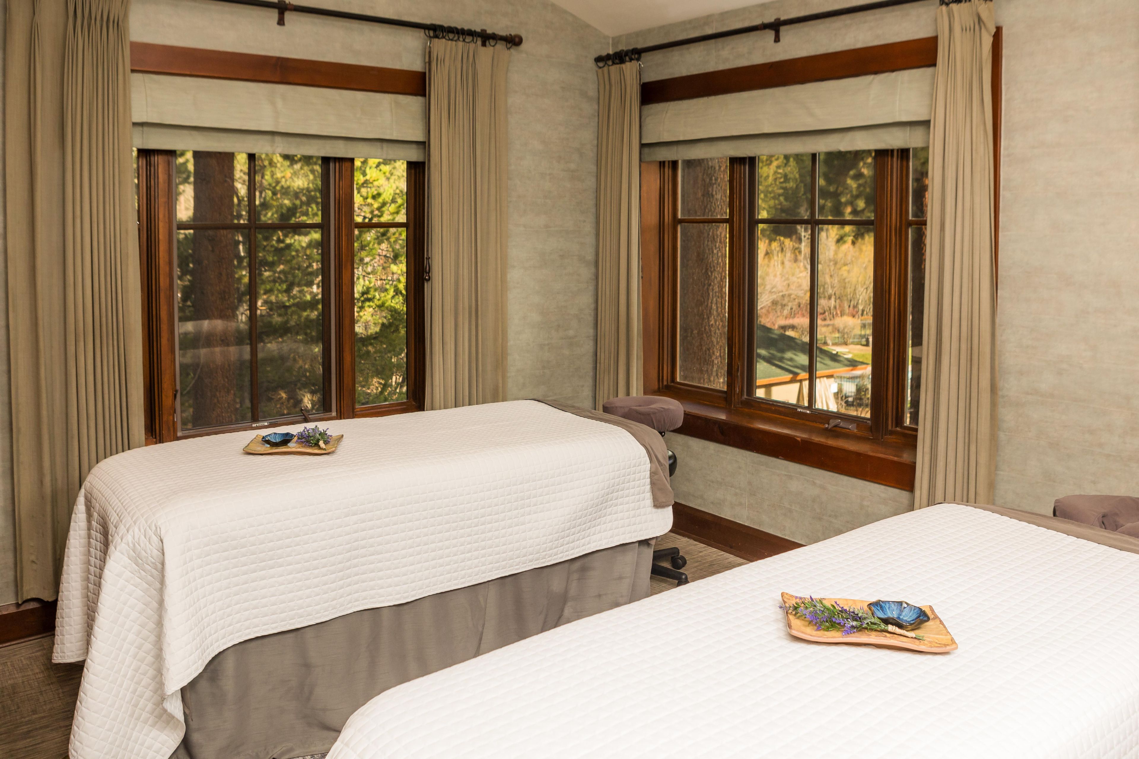 Two single beds with headboards, a wooden bench, and a view of trees through large windows.