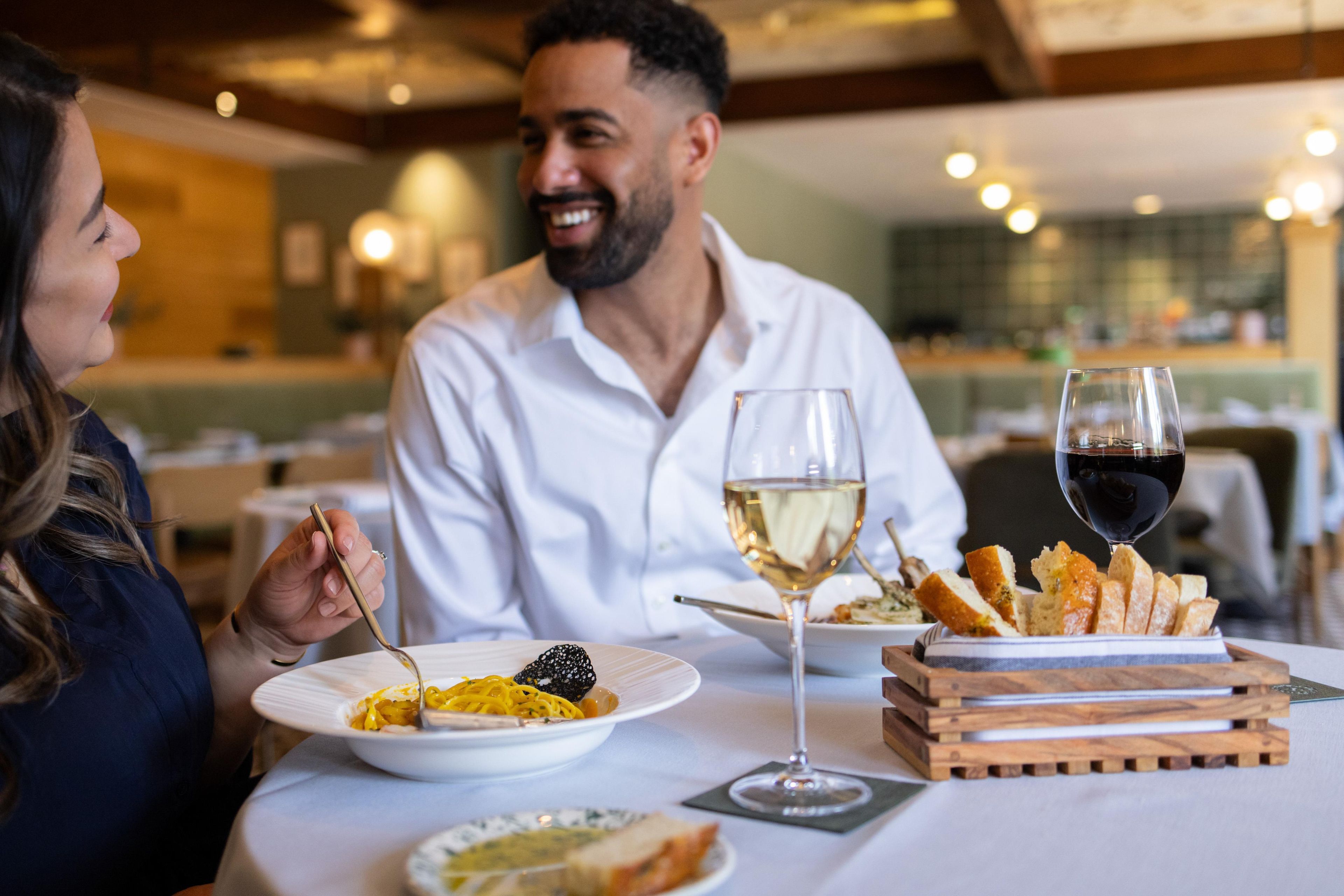 Une table à manger avec des assiettes remplies de nourriture, des verres de vin et une planche en bois sur laquelle est disposé du pain.