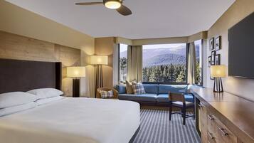 Quarto de hotel com cama, escrivaninha, cadeira, sofá e vista para uma paisagem montanhosa através de amplas janelas.