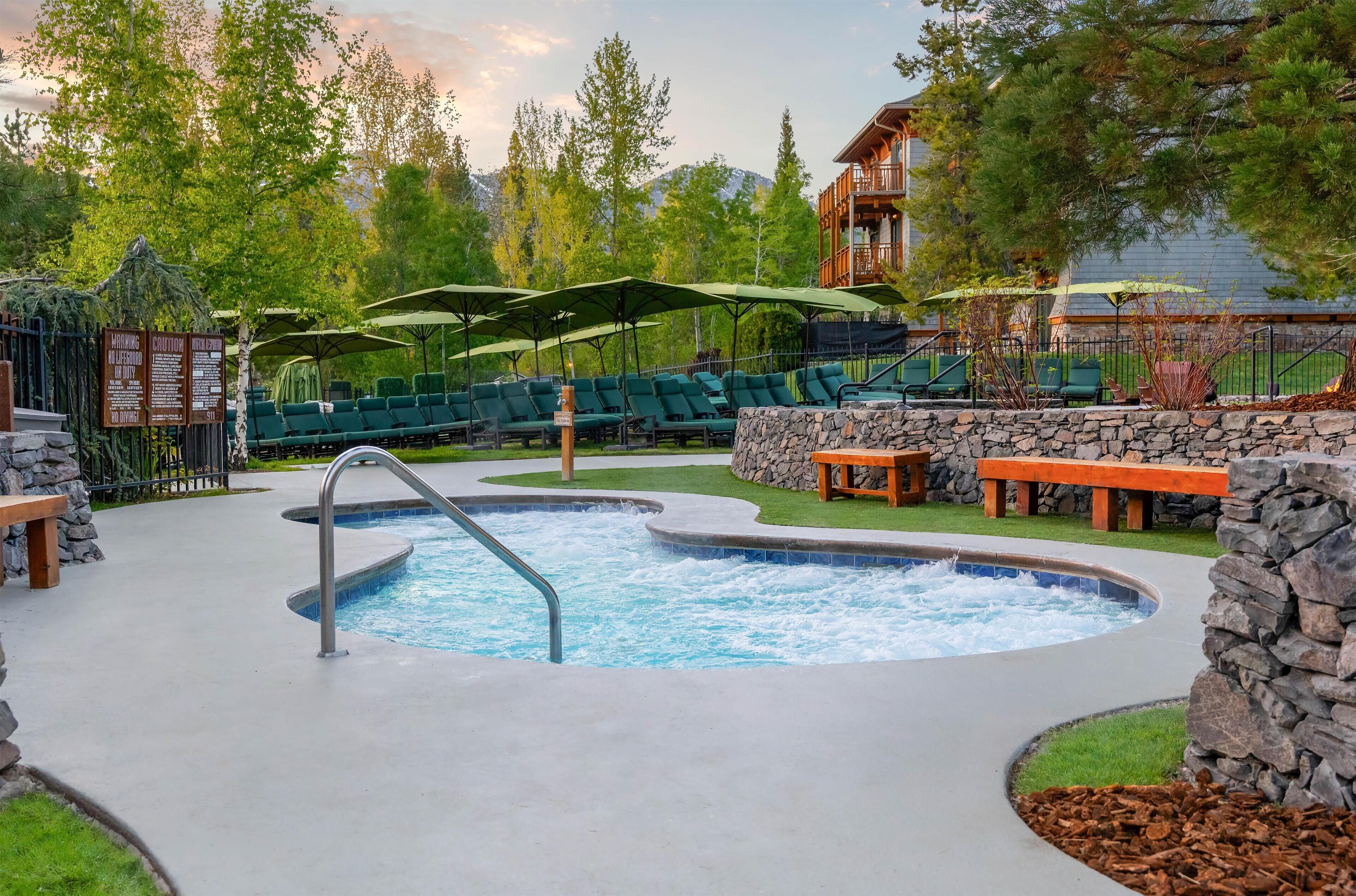 A hot tub surrounded by lounge chairs and umbrellas, with a stone wall and wooden deck.