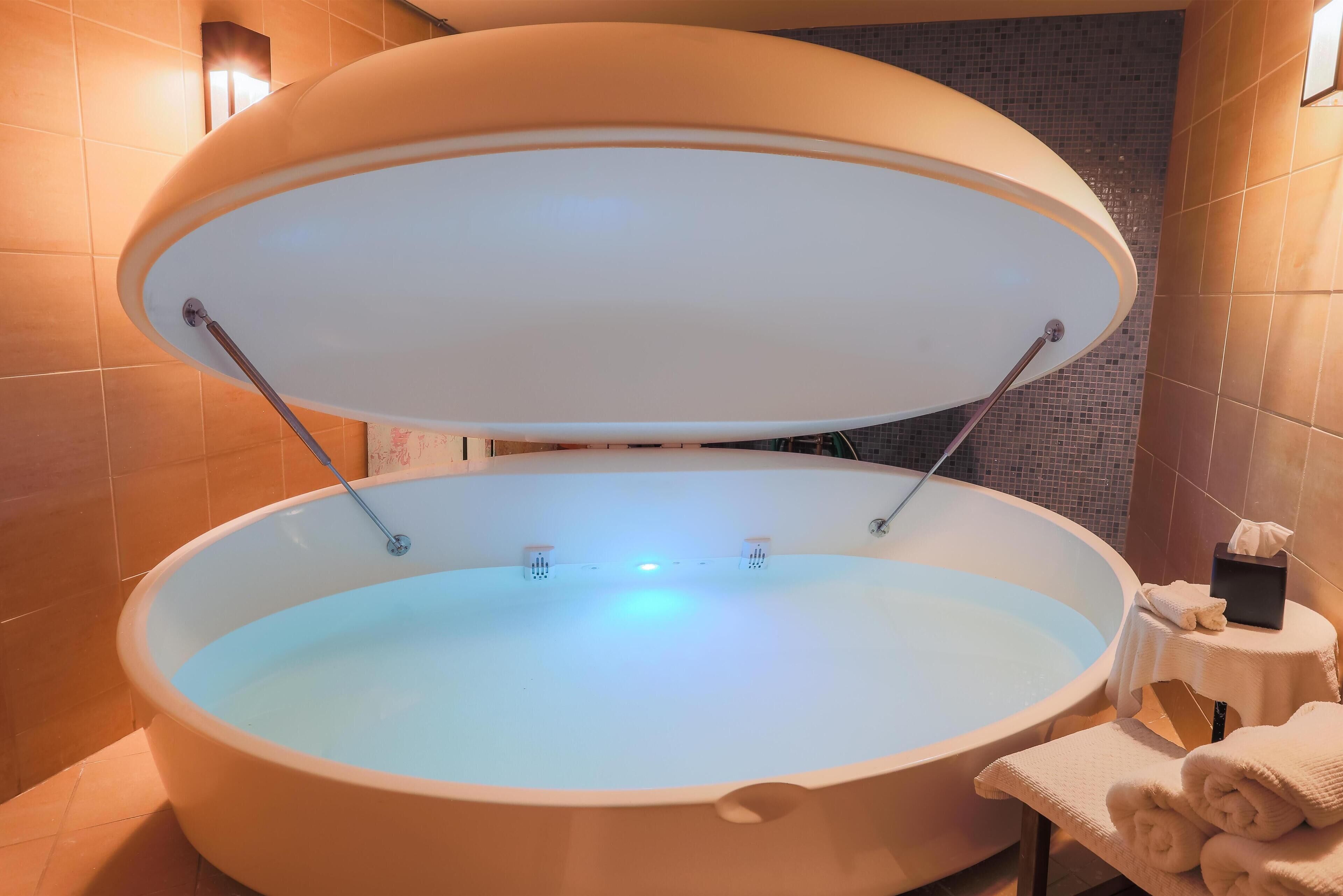 Un grand bain à remous de forme ovale, doté d’un couvercle transparent, installé dans une salle de bain moderne aux murs carrelés, avec une petite table sur laquelle sont posées des serviettes.