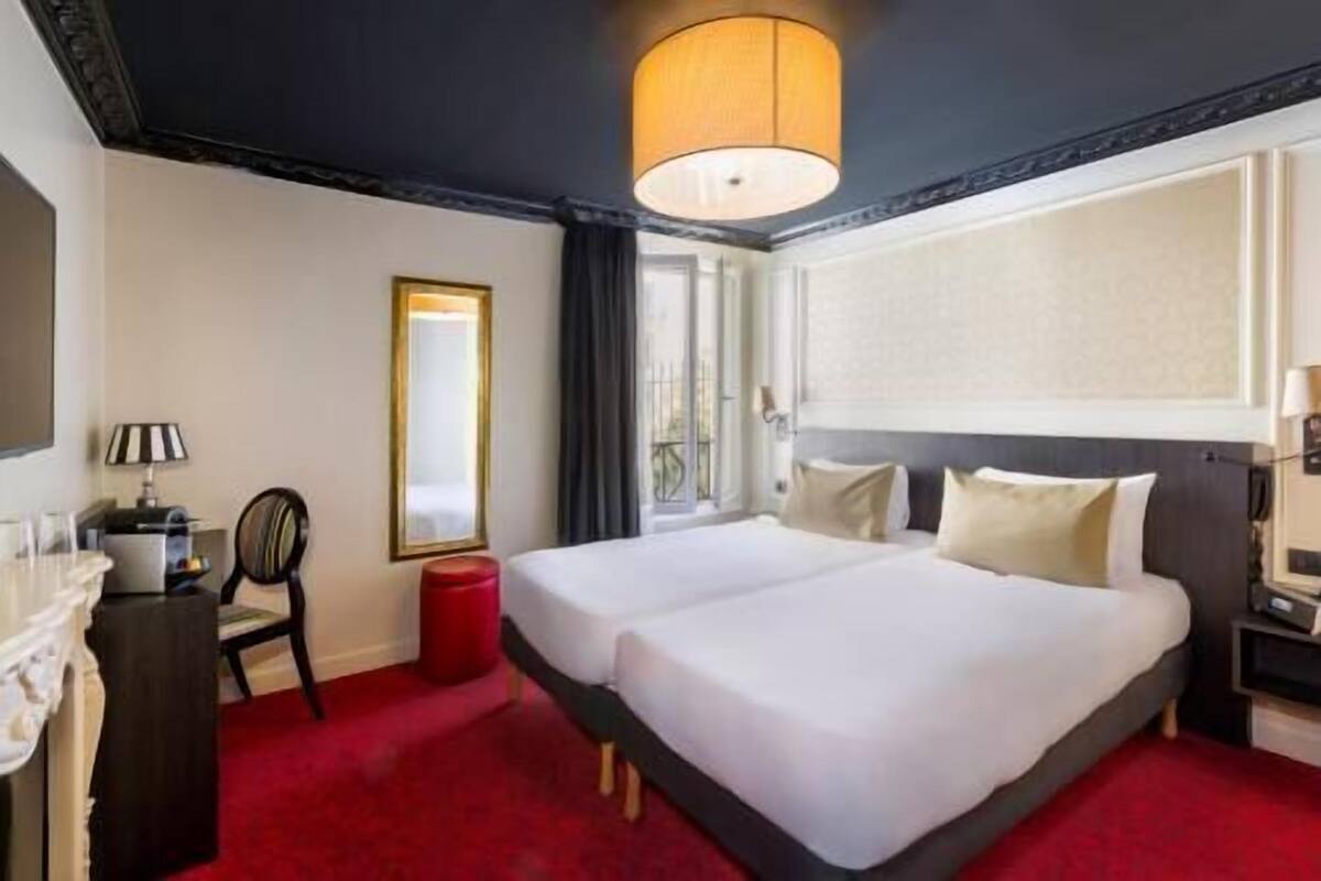 A hotel room with two beds, a desk, a chair, a red ottoman, a lamp, and a large window.