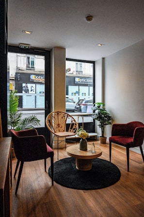 Une pièce avec un plancher en bois, deux chaises, une table avec un vase, et une vue sur un bâtiment avec une enseigne.