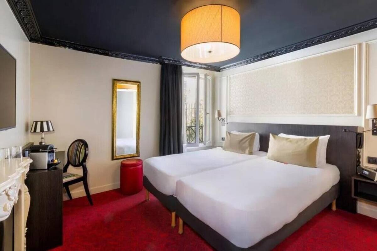 A hotel room with two beds, a red carpet, a large mirror, a desk with a lamp, and a view of the outdoors.