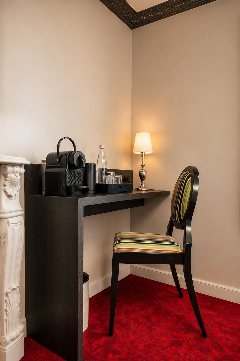 A corner room with a desk, chair, lamp, and a red carpet.