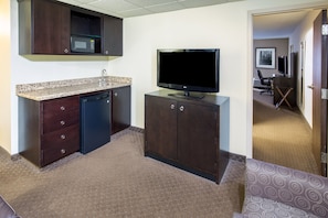 A compact hotel room with a kitchenette, a flat-screen TV, a small desk, and a chair.