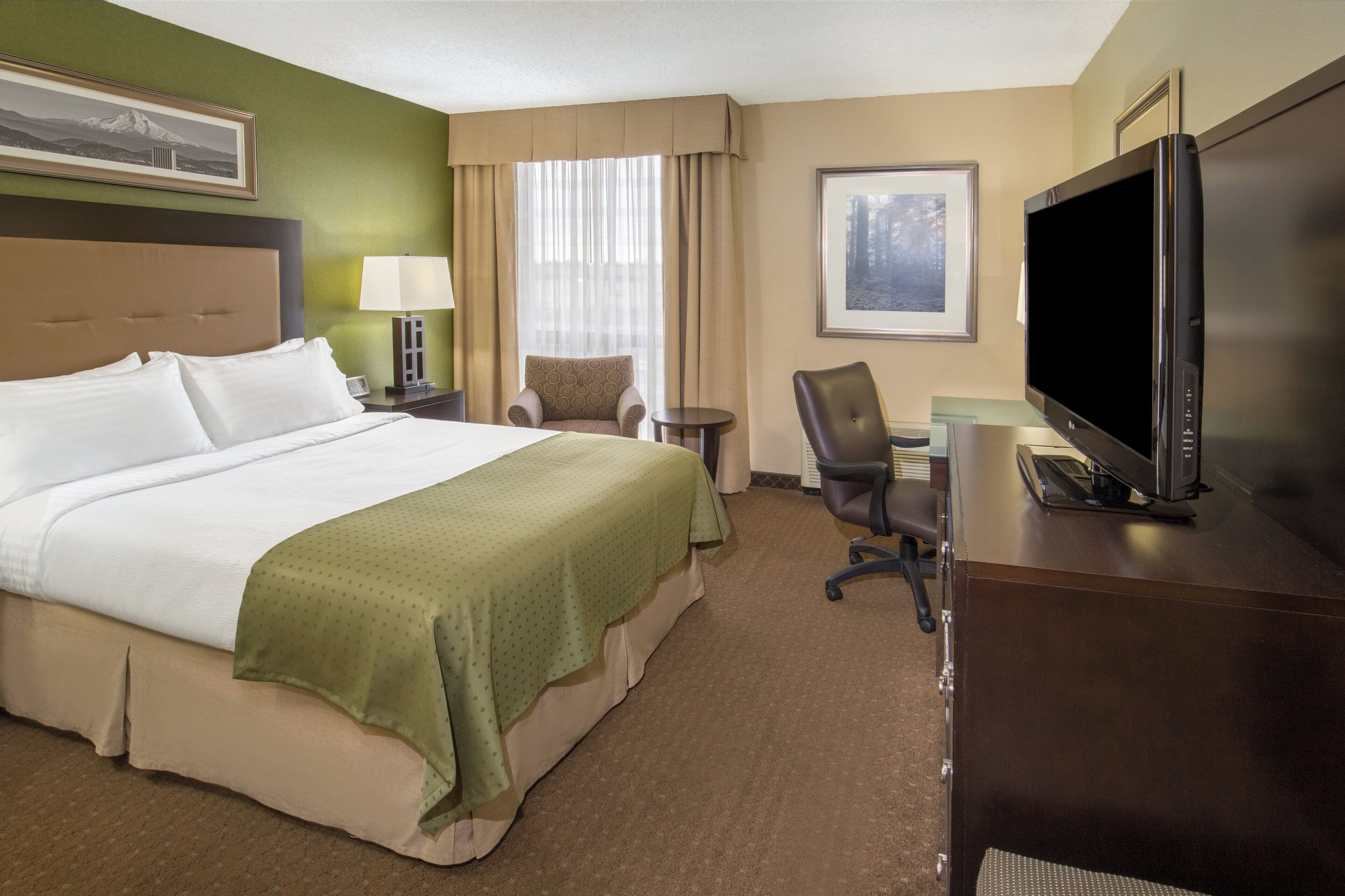 A hotel room with a large bed, a desk with a computer, a TV, a chair, a lamp, and a framed picture on the wall.