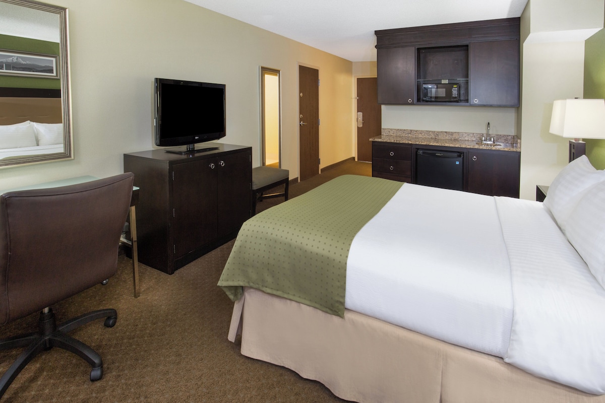A hotel room with a bed, a desk with a television, a chair, a kitchenette, and a bathroom.