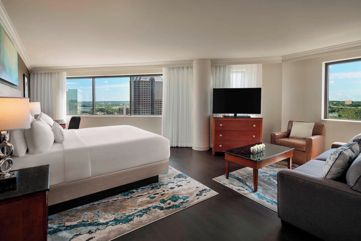 Ein Hotelzimmer mit einem großen Bett, einem Fernseher, einer Sitzecke und Blick auf die Stadt.