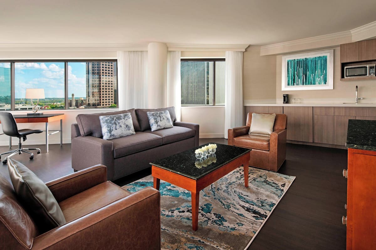 Ein modernes Wohnzimmer mit Sofa, Sessel und Couchtisch, einem Schreibtisch mit Stuhl und Blick auf die Stadt durch große Fenster.