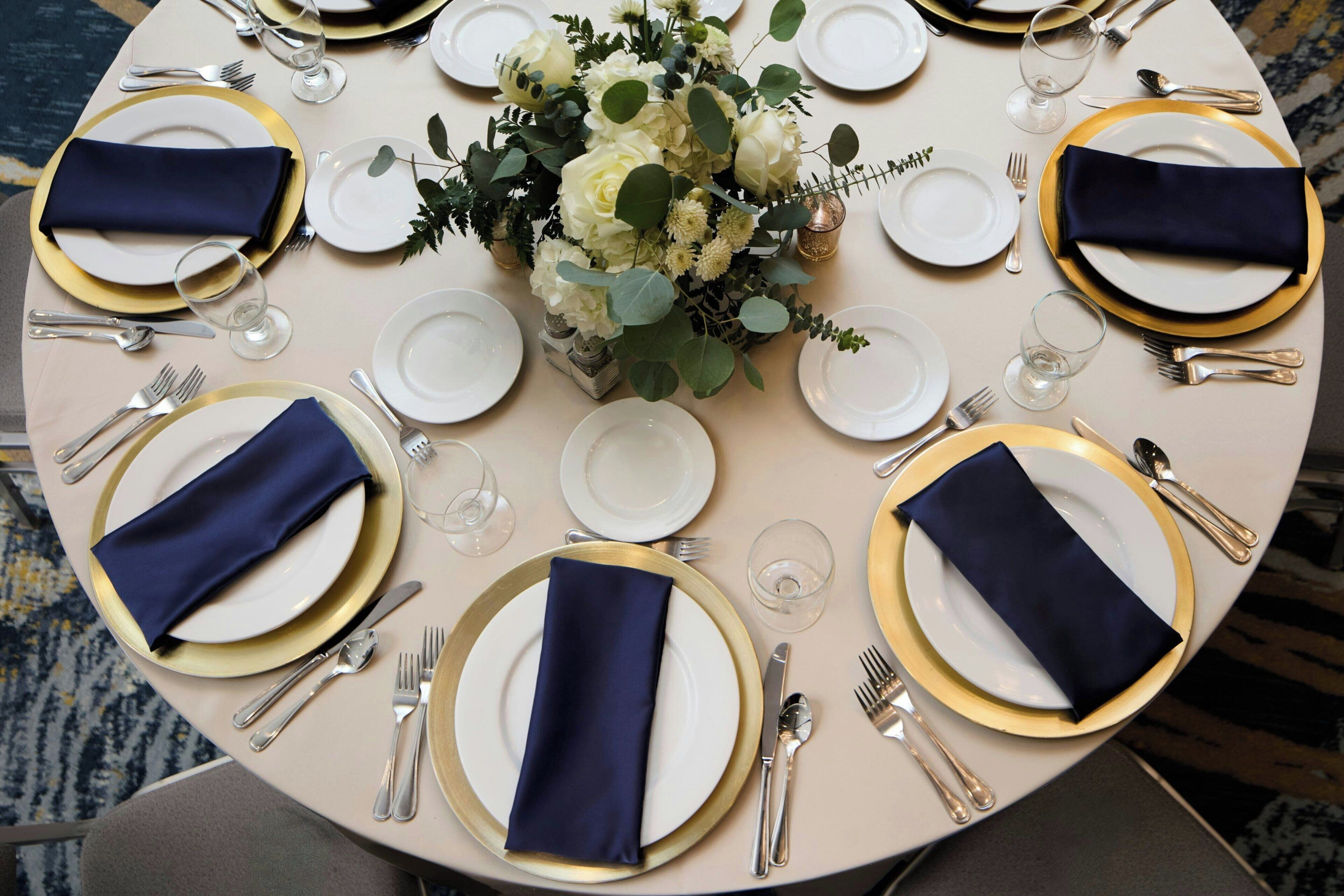 A round table set for a formal dinner with white plates, gold rims, and navy blue napkins.