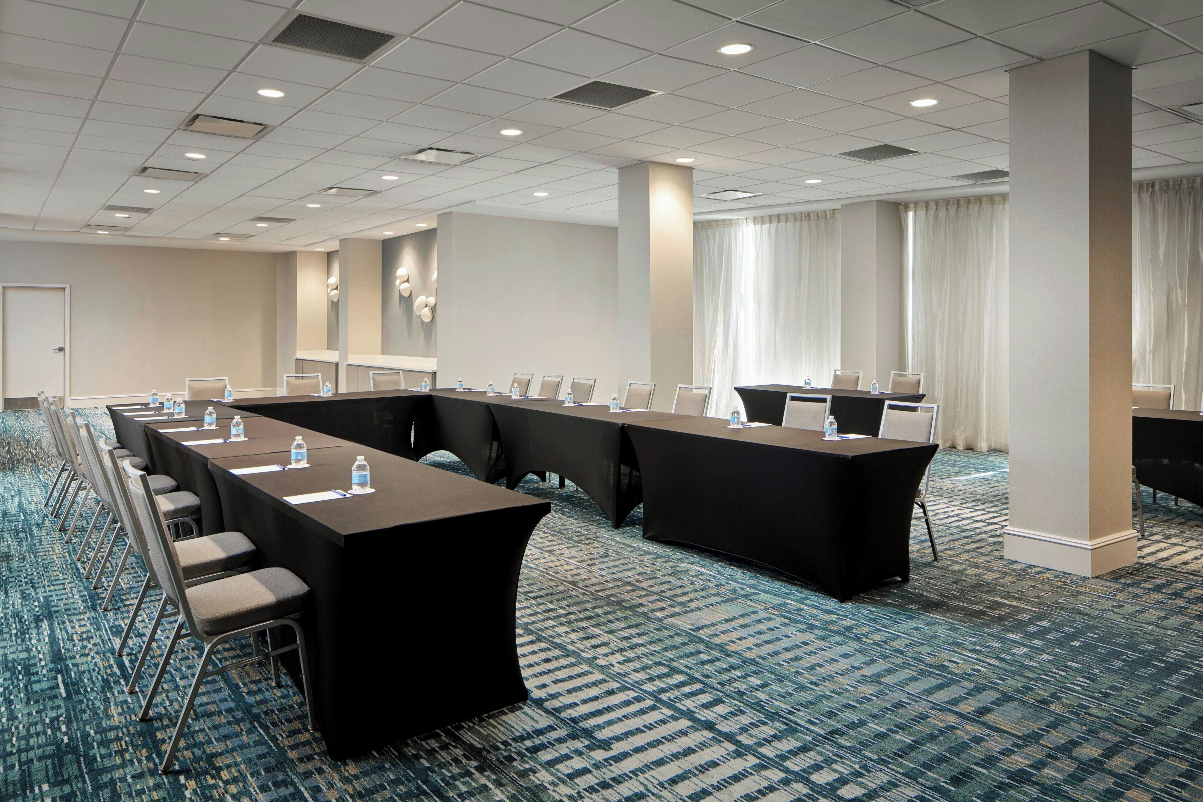 Un salón de conferencias con mesas largas y sillas, piso alfombrado y un gran ventanal con cortinas.